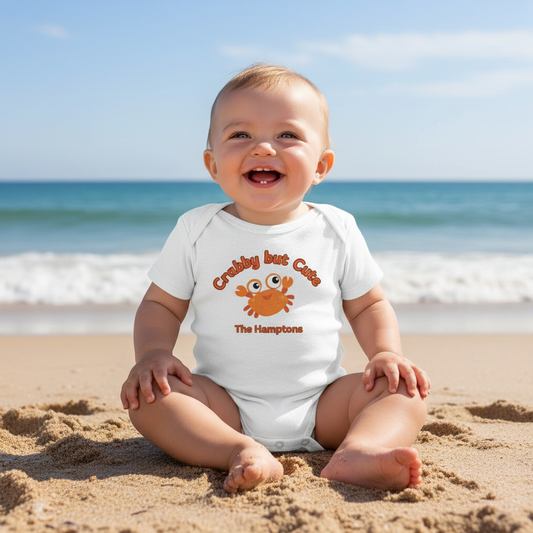 Baby sitting on a beach wearing a white onesie with a crab design and text.