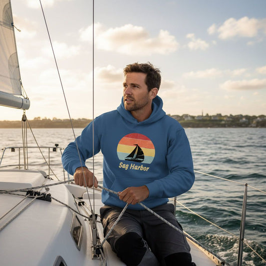 Man sailing in Sag Harbor Unisex Hoodie - Coastal Comfort & Style, featuring retro sailboat graphic, perfect for beach town adventures.