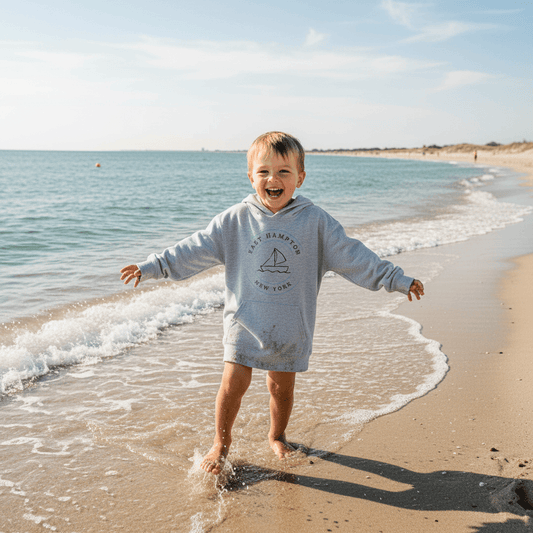 Smiling child wearing Kids East Hampton Sweatshirt in grey, playing on a sunny beach, showcasing cozy coastal style apparel for kids.