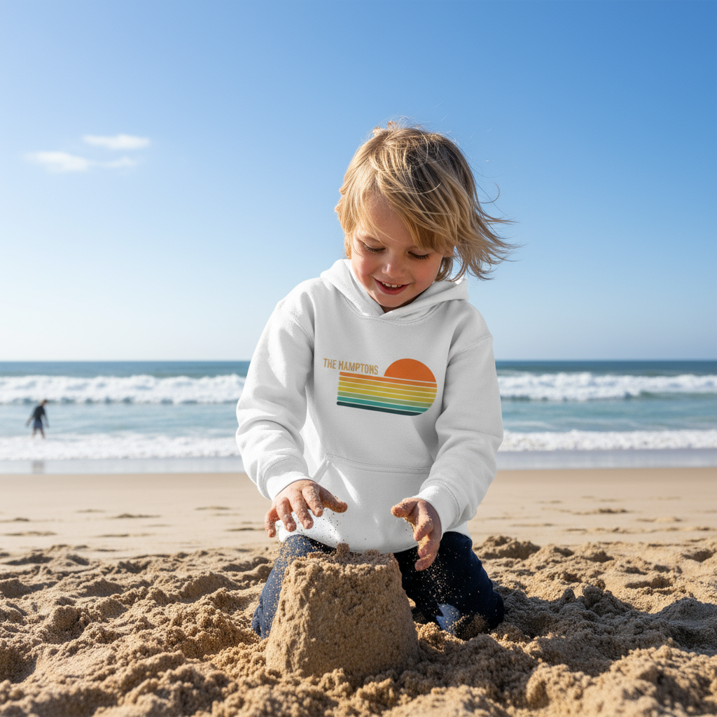 Child building a sandcastle on a beach wearing a white hoodie with a colorful logo.